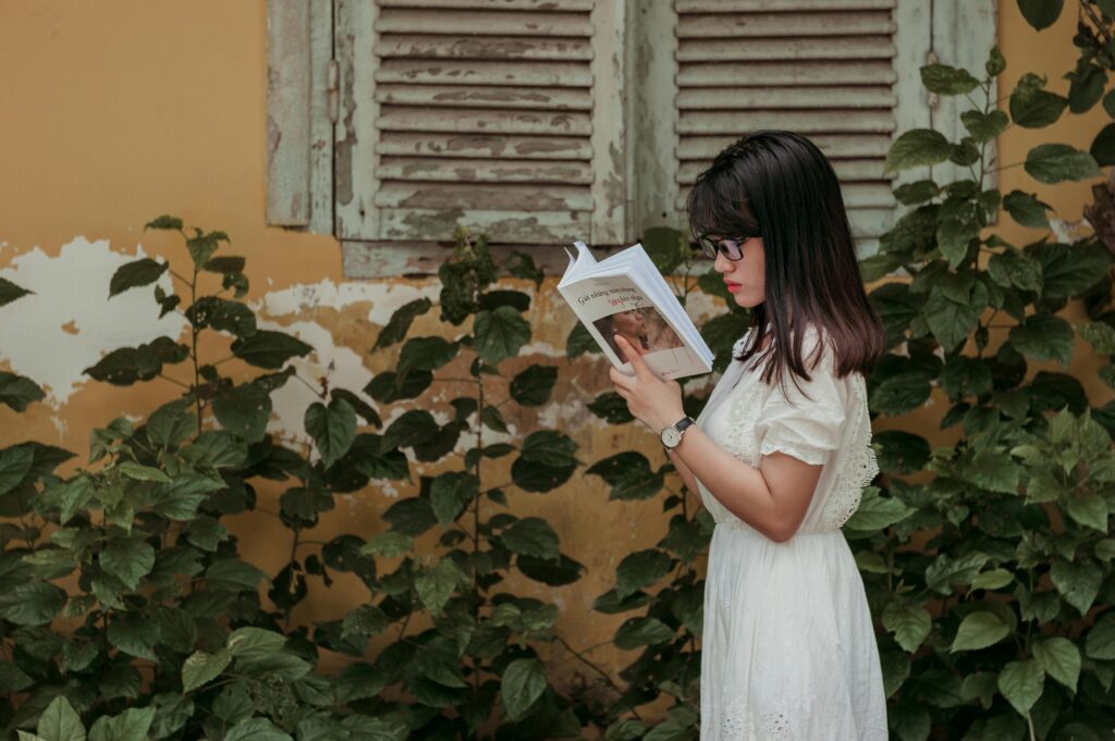 Woman in white dress reading a book outdoors near vintage window shutters and foliage.